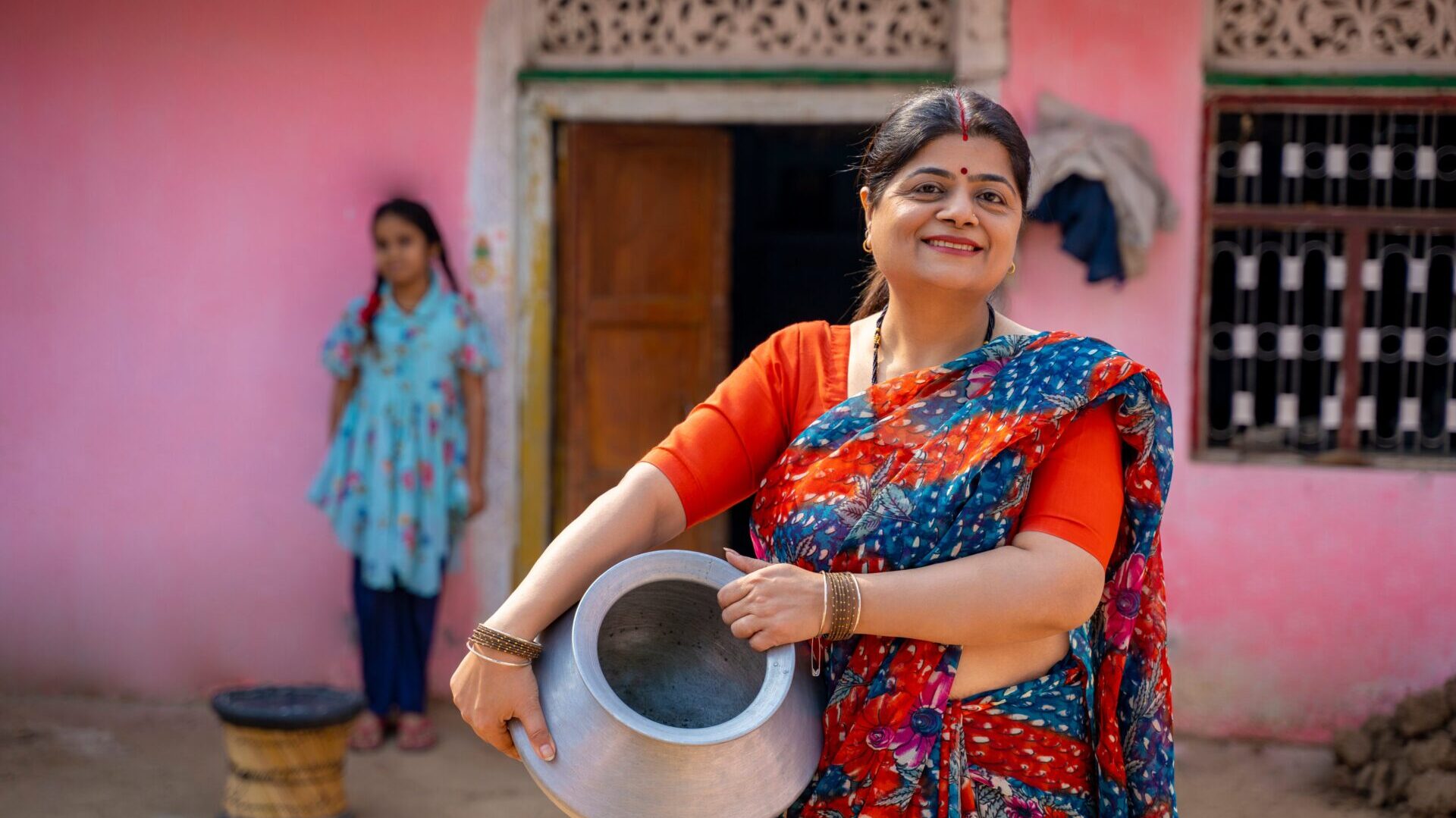 A smiling woman in a vibrant orange and blue saree holding a traditional metal pot, representing the global community of women navigating menopause.