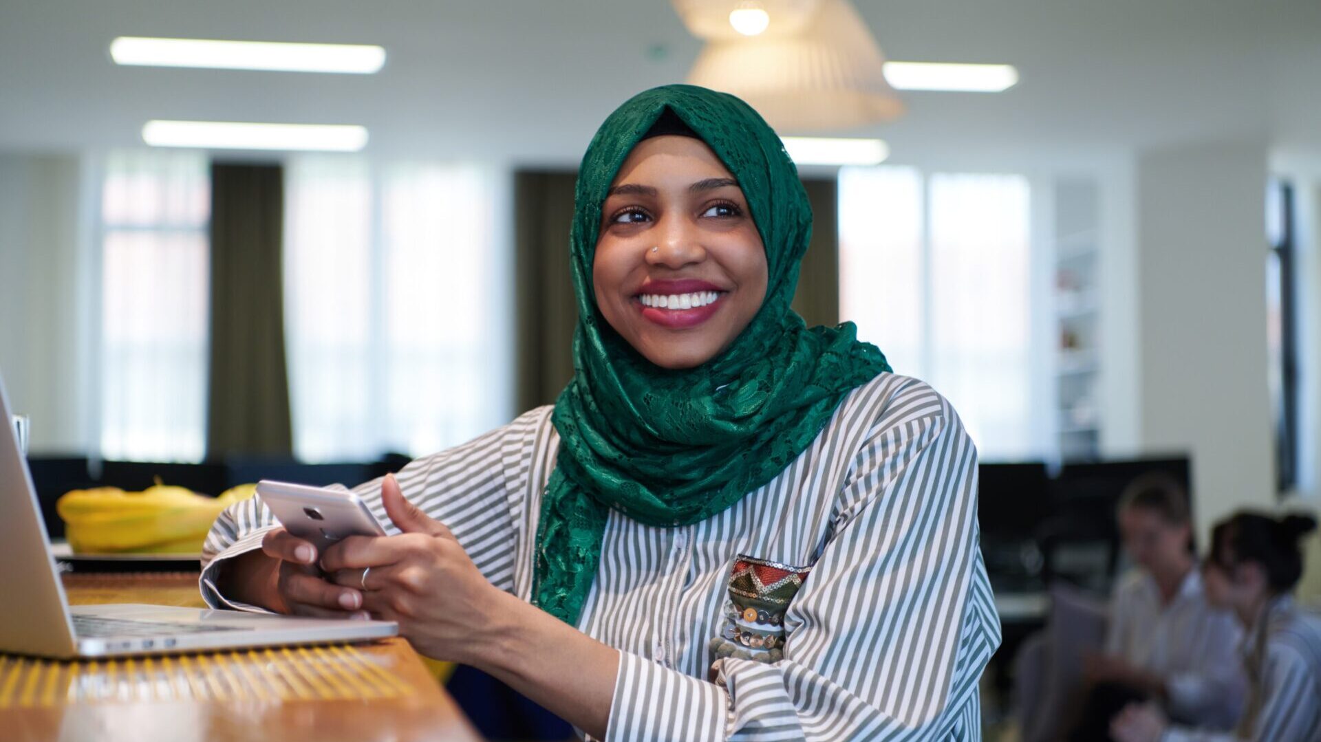 Smiling woman wearing a green hijab sitting in a modern office, holding a smartphone beside a laptop, representing cultural diversity and women in midlife.